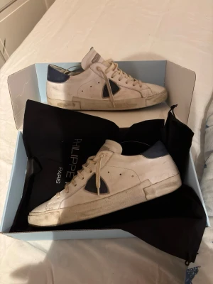 Philippe Model vita sneakers med blå detaljer - Säljer ett par snygga Philippe Model sneakers i vitt skinn med mörkblå detaljer på sidan och hälen. Skorna har rund tå, snörning och platt sula i gummi. Klassisk design med silverfärgad logga på tungan och svart yttersula. Perfekt för dig som gillar stilrena sneakers.