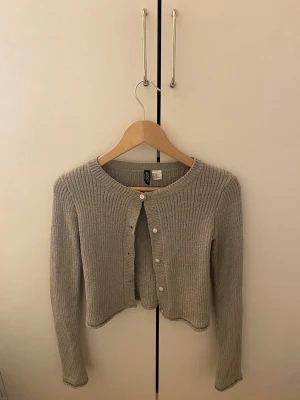 Beige ribbad kofta från HM - Säljer en Grå/grön ribbad kofta från HM med rund halsringning och långa ärmar. Koftan har en croppad passform och stängs med små vita knappar framtill. Perfekt att slänga över en topp för en chill look.