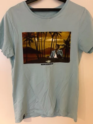 Ljusblå t-shirt med tryck från Dedicated - Ljusblå t-shirt från Dedicated i 100% ekologisk bomull. Framsidan har ett stort tryck med palmer, solnedgång och texten 'Every dog has his day'. Klassisk passform och rund hals. Perfekt för dig som gillar unika prints och streetstyle.