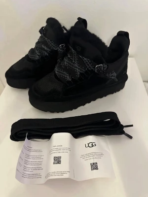 Svarta sneakers från UGG med päls - Säljer ett par chunky svarta sneakers från UGG med fluffig päls på insidan, grov sula och breda snören i svart och grått mönster. Skorna har UGG-logga på både plös och häl, samt detaljer i mocka och mesh. Perfekta för dig som vill ha både stil och komfort.