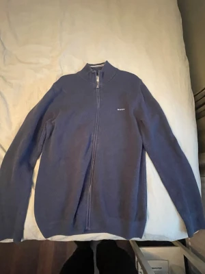 Blå zip-kofta från GANT - Säljer en stilren marinblå kofta från GANT med hel dragkedja och liten logga på bröstet. Tröjan har långärmad design och stårkrage, perfekt för lager-på-lager. Materialet är mjukt och stickat, vilket gör den både bekväm och snygg. Knappt använt!