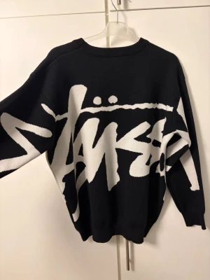 Svart stickad Stüssy tröja med logga - Säljer en svart stickad tröja från Stüssy med stor vit logga över hela framsidan. Tröjan har rund hals, långa ärmar och en loose passform. Perfekt för dig som gillar streetwear och vill sticka ut med en fet grafisk design. STORLEK S/M beroende på hur man vill ha passformen