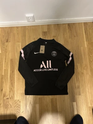 Psg track (överdel) - Paris Saint-Germain träningströja från Nike i svart med ljusrosa detaljer och PSG-logga på bröstet. Tröjan har lång ärm, halv dragkedja och tryck på både fram- och baksida. Materialet är polyester och designen är sportig med snygga kontraster. Priset är bara för överdelen!