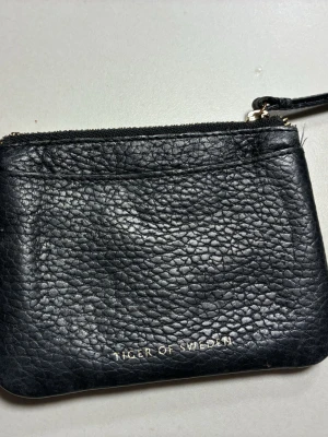 korthållare  från Tiger of Sweden - Snygg svart korthållare från Tiger of Sweden i äkta skinn med textur. Plånboken har både dragkedja och ett fack med tryckknapp, perfekt för kort och smågrejer. Stilren design med guldfärgade detaljer och logga framtill.