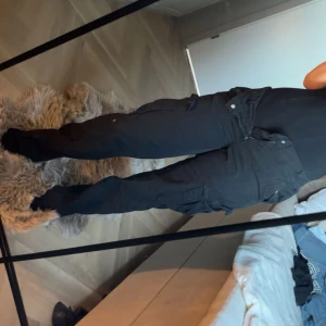 Svarta cargopants med raka ben - Säljer ett par svarta cargopants med raka ben och flera praktiska fickor på sidorna. Byxorna är lowaist och är tillverkade i ett slitstarkt bomullsmaterial.