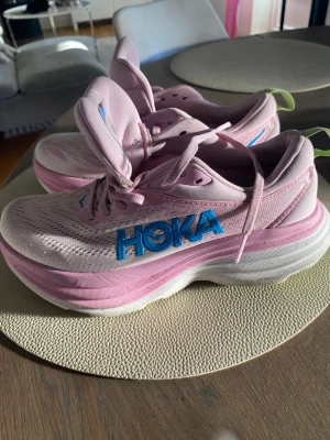 Rosa Hoka sneakers med blå logga - Hoka Bondi 8 - Säljer ett par snygga rosa Hoka sneakers med blå logga på sidan och vit, tjock sula med limegröna detaljer undertill.  Knappt använda då modellen inte passade så bra.