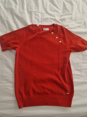 Orange stickad tröja Holly & Whyte - Snygg orange stickad tröja från Holly & Whyte by Lindex i storlek M. Tröjan har korta ärmar, rund hals och tre dekorativa knappar vid axeln. Ribbad kant vid hals, ärmslut och nederkant. Perfekt för dig som gillar stilrena detaljer. Den ser röd ut men den är orange!!