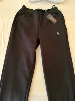 Svarta mjukisbyxor Polo Ralph Lauren - Svarta mjukisbyxor från Polo Ralph Lauren med klassisk broderad logga på benet. Byxorna har ribbad midja med snörning och sidofickor. Materialet är mjukt och skönt, perfekt för chill eller träning. Snygg och enkel design som funkar till allt. Passar inte på fullvuxna män! 