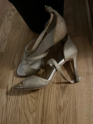 Glittriga guldiga sandaler med klack - Säljer ett par glittriga sandaler från Catwalk i guld med öppen tå och smala remmar som korsar foten. Skorna har hög klack och spänne vid ankeln. Perfekta för dig som vill sticka ut med extra bling på fötterna.