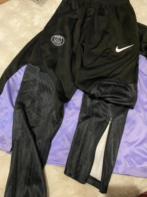 Svarta PSG Nike träningsbyxor - Svarta träningsbyxor från Nike med PSG-logga på höften och vit Nike-swoosh. Byxorna har subtilt mönster längs benen, dragkedjor vid bensluten och är gjorda i ett syntetmaterial som passar perfekt för fotbollsträning.