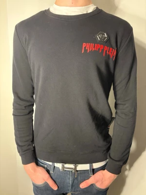 Svart Philipp Plein sweatshirt - Svart sweatshirt från Philipp Plein med rött tryck och logga på bröstet. Tröjan har rund hals, långa ärmar och en normal passform. Materialet är mjuk bomull som känns skön mot huden. Perfekt för dig som gillar streetwear och vill sticka ut med en snygg designertröja.