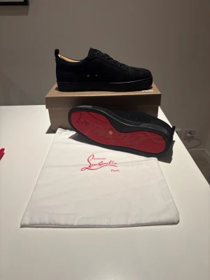 Svarta Sneakers från Christian Louboutin - Snygga svarta sneakers från Christian Louboutin med klassisk röd sula och rund tå. Skorna har snörning och är tillverkade i mocka med mjukt beige foder. Perfekt för dig som vill ha en exklusiv och stilren look med en ikonisk detalj under foten. Kan ej garantera äkthet!