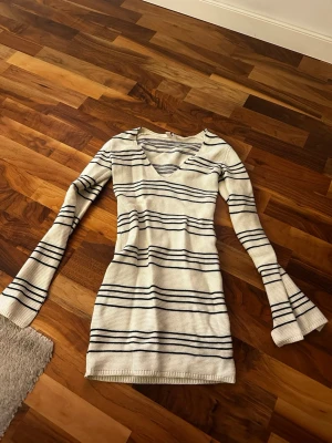 Randig vit stickad kortklänning - Säljer en vit stickad kortklänning med svarta horisontella ränder. Klänningen har långärmade ärmar med lätt utsvängd form och en snygg v-ringad hals. 