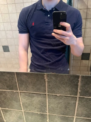 Marinblå pikétröja från Ralph Lauren - Klassisk marinblå pikétröja från Ralph Lauren med röd broderad logga på bröstet. Tröjan har krage, två knappar vid halsen och korta ärmar. Tillverkad i mjuk bomull som känns skön mot huden. Perfekt till jeans eller shorts för en clean look. Vid frågor, tveka inte på att skicka