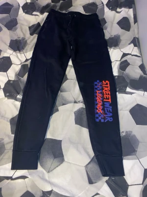 Svarta mjukisbyxor Primark Streetwear - Svarta mjukisbyxor från Primark med trycket 'STREETWEAR LEGENDS' i orange och blått på ena benet. Byxorna har resår i midjan och muddar vid bensluten. Sköna och casual med streetstyle-vibe, perfekta för chill eller häng.