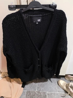Svart stickad kofta från 157 - Säljer en svart stickad kofta från 157 i storlek M. Koftan har grovstickad struktur, v-ringning, knappar framtill och två fickor. Perfekt att slänga över en t-shirt för en chill look. Passar dig som gillar loose fit och enkel stil.