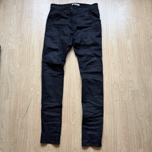 NostraSantissima vaxade jeans - Svarta vaxade jeans från NostraSantissima i slim fit  och flera fickor fram och bak. Byxorna har knapp och dragkedja i midjan samt bälteshällor. Materialet är 97% bomull och 3% elastan vilket ger en skön stretch. Snyggt minimalistiskt utseende och praktiska fickor. Knappt använda. Storlek 46 (stora i storlek). Midja mäter 43 cm 