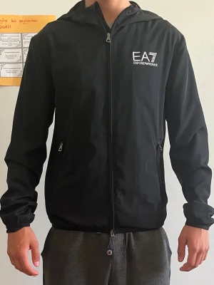 Svart vindjacka EA7 Emporio Armani - Svart vindjacka från EA7 Emporio Armani med huva och dragkedja framtill. Jackan har två sidofickor med dragkedja och vit logotyp på bröstet. Lätt och smidig design, perfekt för dig som gillar stilrena plagg med sportig vibe.