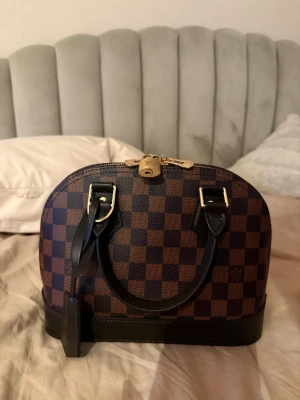 Louis Vuitton Damier Alma handväska - Säljer en ikonisk Louis Vuitton Alma handväska i Damier-mönster. Väskan är brun och mörkbrun med guldfärgade detaljer, lås och nyckel. Insidan är klädd i rött tyg och har en praktisk ficka. Handtag och detaljer är i svart skinn. Perfekt för dig som gillar lyxiga accessoarer.