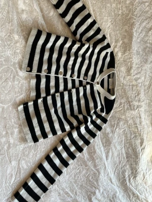 Randig kofta från Zara i svart och vitt - Säljer en ribbstickad kofta från Zara i storlek 152. Koftan har breda svarta och vita ränder, rund hals och silvriga knappar framtill. Materialet är stickat och modellen är normal passform med långa ärmar. Perfekt för dig som gillar klassisk stil med en twist.