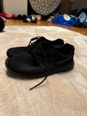 Svarta Nike sneakers med snörning - Säljer ett par helsvarta Nike sneakers med klassisk Swoosh-logga på sidan. Skorna har rund tå, platt sula och är tillverkade i ett ventilerande syntetmaterial med mesh-ytor. Perfekta för dig som gillar stilrena och sportiga skor med skön passform.