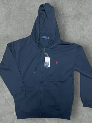 Ralph Lauren Polo Zip – Blå - Stilren och tidlös hoodie från Ralph Lauren. Blå färg med ikonisk Polo-logga på bröstet. Bekväm passform och hög kvalitet som håller länge. Perfekt för både casual och mer uppklädda outfits. I fint skick. 
