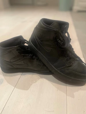 Svarta Nike Jordan 1 Highs strl 41 - Säljer ett par svarta Nike Jordan 1 Highs i storlek 41. Skorna har en klassisk high-top siluett med snörning och rund tå. Tillverkade i skinn med detaljerade sömmar och Nike-logga på sidan. Perfekta för dig som gillar streetwear och vill ha en ikonisk sneaker.