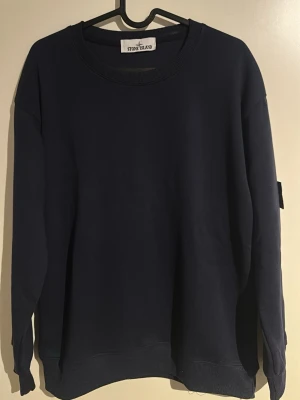 Mörkblå sweatshirt från Stone Island - Säljer en mörkblå sweatshirt från Stone Island med klassisk rund hals och ribbade muddar. Tröjan har den ikoniska Stone Island-patchen på ärmen och är gjord i mjuk bomull. Perfekt för dig som gillar streetwear och snygga detaljer.