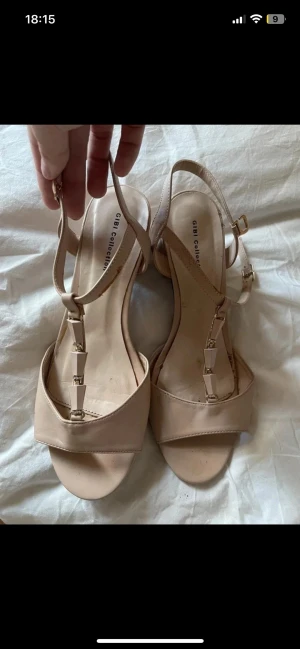 Beige sandaler med klack från Gibi - Snygga beige sandaler från Gibi Collection med bred rem över tårna och dekorativa detaljer i guld på remmen. Skorna har en stabil, låg klack och justerbart spänne vid ankeln. Materialet är syntet och designen är stilren med öppen tå. finns märken på den se på bilderna