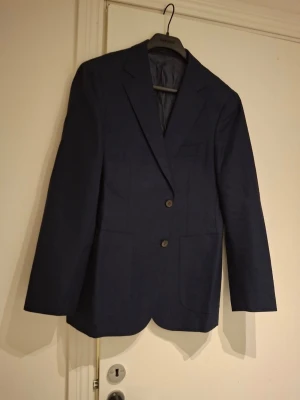 Mörkblå kavaj med knappar - Snygg mörkblå kavaj med klassisk passform och två knappar framtill. Kavajen har tre dekorativa knappar vid ärmslutet och är helfodrad. Perfekt för dig som vill ha en stilren och tidlös look. Materialet känns mjukt och har en lätt struktur. Storlek 24? 