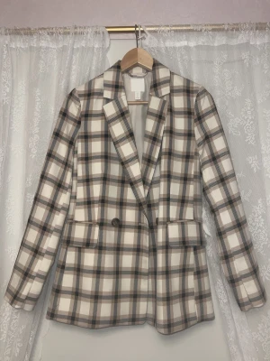 Rutig kavaj från H&M i beige och svart - Snygg rutig kavaj från H&M i beige, svart och vitt. Klassisk modell med långa ärmar, två knappar framtill och fickor med lock. Perfekt för dig som vill ha en stilren och trendig look. Tidlös design som funkar till många outfits.