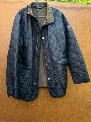 Svart quiltad jacka från Gant - Quiltad svart jacka från Gant med brunt foder och hög krage. Jackan har stora fickor framtill, tryckknappar och dragkedja. Klassisk badge på ärmen och snyggt rutmönster över hela jackan. Perfekt för kyliga dagar.