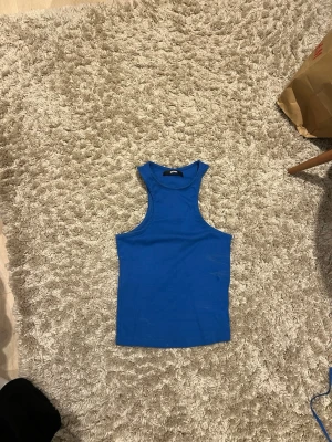 Blå ribbad linnetopp från Gina Tricot - Säljer en enkel och snygg blå linnetopp från Gina Tricot. Toppen är ribbad och ärmlös med rund halsringning och tight passform. Perfekt att styla till jeans eller kjol för en clean look.