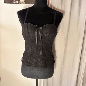 Svart spetskorsett med rosett - Säljer en svart korsett med tunna axelband och snygg spetsdetalj över hela toppen. Framtill finns en liten rosett som ger en extra touch. Modellen är tight och har en feminin siluett med blommigt spetsmönster.