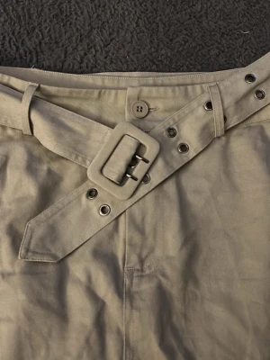 Beige kortkjol med bälte från Gina Tricot - Säljer en beige kortkjol från Gina Tricot i storlek L. Kjolen har ett brett bälte med metallhål och stor spänne, samt två stora fickor framtill. Materialet är bomull och kjolen har en cool utility-vibe som passar till sneakers eller boots. Helt ny men utan prislapp 