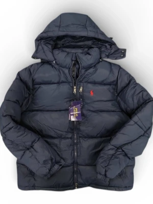 Mörkblå pufferjacka från Polo Ralph Lauren - Snygg mörkblå pufferjacka från Polo Ralph Lauren med röd broderad logga på bröstet. Jackan har huva, dragkedja framtill och quiltad design. Perfekt för kalla dagar och har en modern, loose passform.