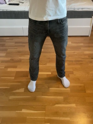 Jeans med slitningar  - Säljer ett par mörkgrå jeans från Jack & Jones med slim passform. Jeansen har klassisk femficksdesign, slitningar och hål på ena benet för en trendig look. Tillverkade i jeansmaterial med dragkedjegylf och bälteshällor.