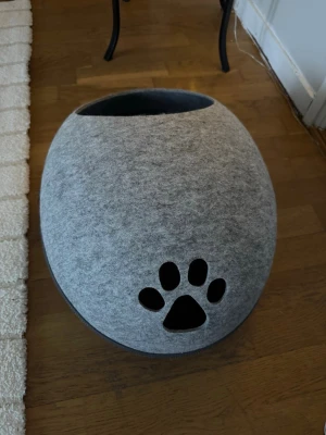 Grå kattkoja i filtmaterial - Säljer en rund och mysig kattkoja i ljusgrå filt med svart tassformad öppning framtill. Koja har dragkedja runt om och en stor öppning upptill för enkel åtkomst. Perfekt för din katt att chilla i.