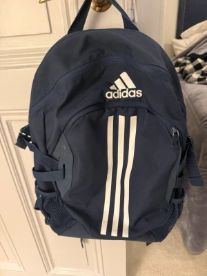 Mörkblå Adidas ryggsäck med vita ränder - Säljer en snygg mörkblå ryggsäck från Adidas med klassiska vita ränder på framsidan och logga både fram och bak. Väskan har flera fack, meshfickor på sidorna och justerbara axelremmar. Perfekt för skola eller träning!