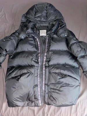 Svart Moncler pufferjacka med huva - Säljer en svart pufferjacka från Moncler med stor huva och feta dragkedjor. Jackan har quiltad design, två sidofickor med dragkedja och Moncler-logga på ärmen. Materialet är glansig polyester och dunfyllning, perfekt för kalla dagar.