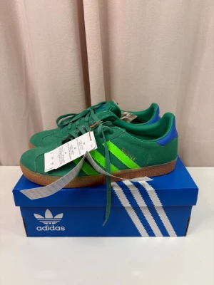 Adidas Gazelle gröna sneakers mocka - Säljer ett par gröna Adidas Gazelle sneakers i mocka med limegröna ränder och blå detalj vid hälen. Skorna har klassisk gummisula och snörning framtill. Perfekta för dig som vill sticka ut med färg och retrostil.