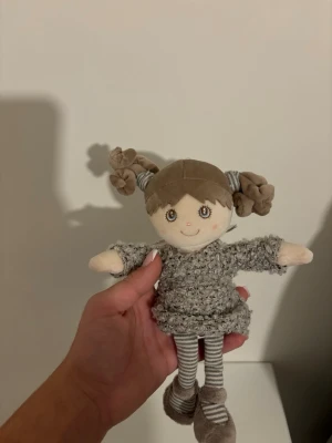Grå stickad väst till docka - Säljer en söt grå stickad väst till docka, med grovstickat mönster och lång ärm. Perfekt för att ge dockan en mysig och trendig look.