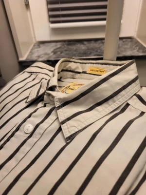 Vintage randig skjorta från Pierre Hector XL 42 - Vit skjorta med svarta vertikala ränder från Pierre Hector Young Collection. Skjortan är i 100% bomull, har klassisk krage, knäppning framtill och en bröstficka med broderad detalj i rött, blått och gult. Perfekt för dig som gillar retrostil.