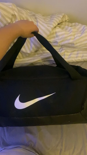 Svart träningsväska från Nike - Säljer en svart träningsväska från Nike med klassisk vit swoosh-logga på sidan. Väskan har rektangulär form, två rejäla handtag och en dragkedja på sidan för extra förvaring. Perfekt storlek för gymmet eller sportaktiviteter.