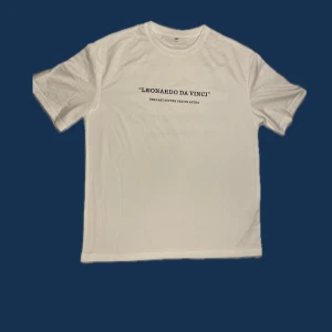Vit t-shirt med Leonardo Da Vinci-tryck - Vit t-shirt med svart texttryck på bröstet där det står 'LEONARDO DA VINCI' och 'THE LAST SUPPER GENIUS ARTIST'. Klassisk rund hals och korta ärmar. Enkel och stilren design, perfekt för dig som gillar konst och minimalism.