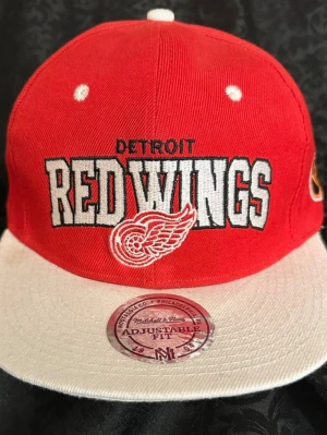 Röd och vit Detroit Red Wings keps - Snygg snapback-keps från Mitchell & Ness med Detroit Red Wings broderat framtill. Kepsen är röd med vit skärm och vita detaljer, justerbar baktill. Materialet är 100% ull och insidan har märkets logga. Perfekt för dig som gillar hockey och vill ha en cool accessoar.