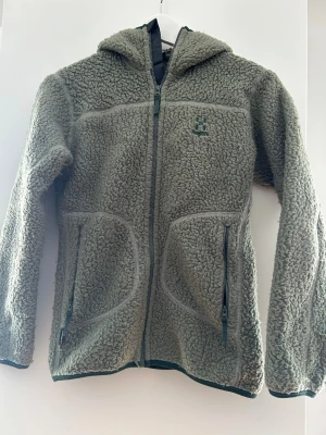 Grön teddyjacka med huva från Haglöfs - Mysig grön teddyjacka från Haglöfs i storlek 152 Junior. Jackan har huva, två fickor med dragkedja och hel dragkedja framtill. Tillverkad i fluffigt fleece-material som håller dig varm under kyliga dagar. Perfekt för dig som gillar outdoor och chill stil.
