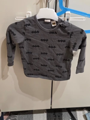 Grå Batman långärmad topp H&M - Säljer en grå långärmad topp från H&M i storlek 80, med svart Batman-logga mönster över hela plagget. Toppen är tillverkad i 100% bomull och har rund hals. Perfekt för små superhjältar som gillar coola tryck.