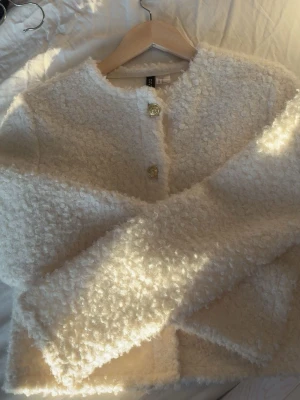 Vit teddyjacka från Divided XS - Säljer en fluffig vit kofta från Divided i storlek XS. Koftan har rund hals, långärmade ärmar och snygga guldfärgade knappar framtill. Materialet är mjukt och ger en cool, cozy vibe. Perfekt för dig som gillar stilrena och trendiga plagg.