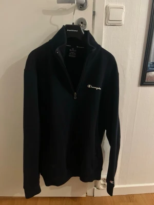 Svart half zip tröja från Champion - Svart långärmad tröja från Champion med half zip och hög krage. Diskret Champion-logga broderad på bröstet och en liten logga på ärmen. Tröjan är i mjukt material och har en clean, sportig look som funkar till chill och streetwear.